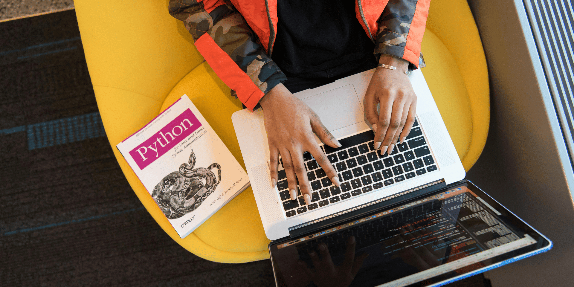 A woman learning to code with Python, typing on a laptop with a Python programming book beside her on a yellow chair